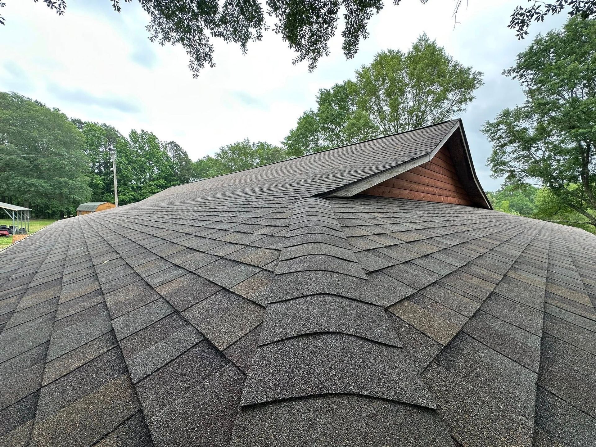 Low angle view of a gray asphalt shingle roof with a wooden gable against a backdrop of trees.