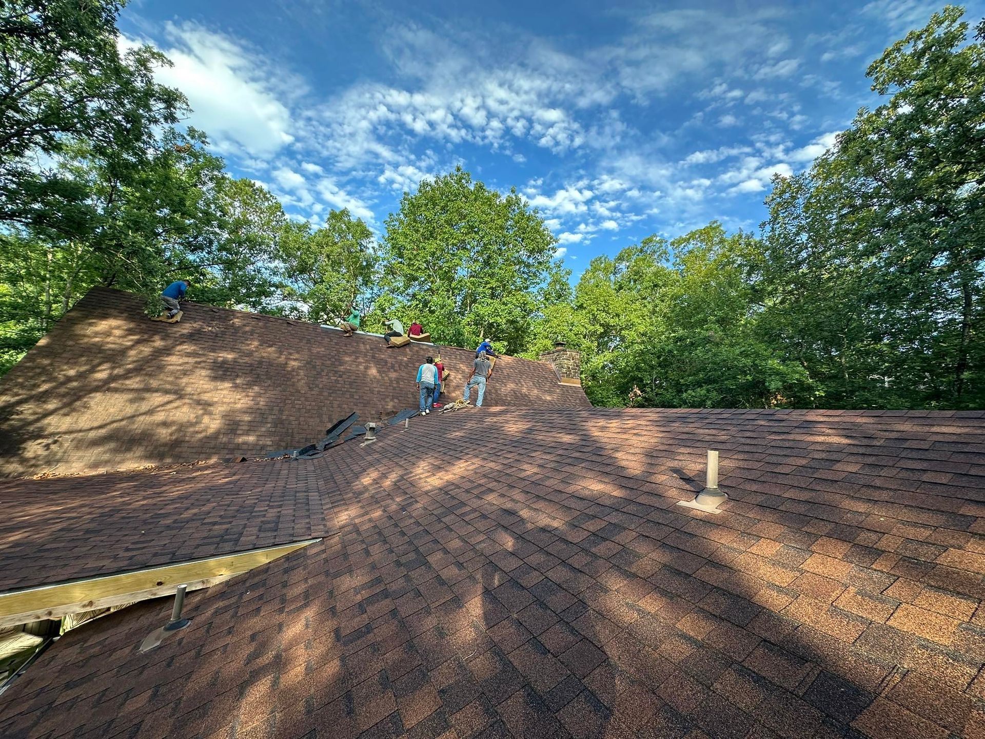 Workers repairing a brown shingle roof on a sunny day, surrounded by green trees and blue sky.