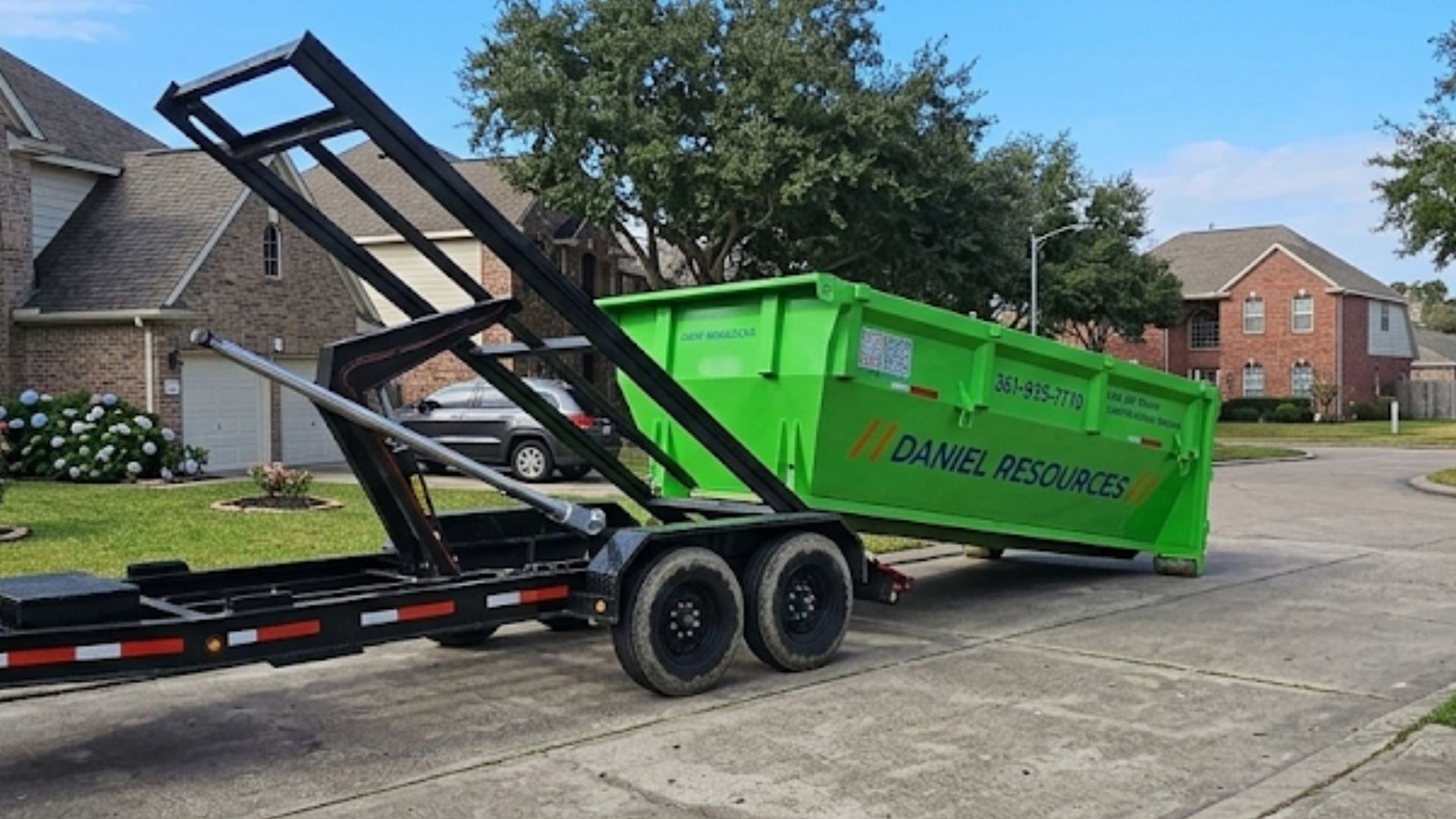 Green dumpster trailer with raised front, parked on a residential street near houses.