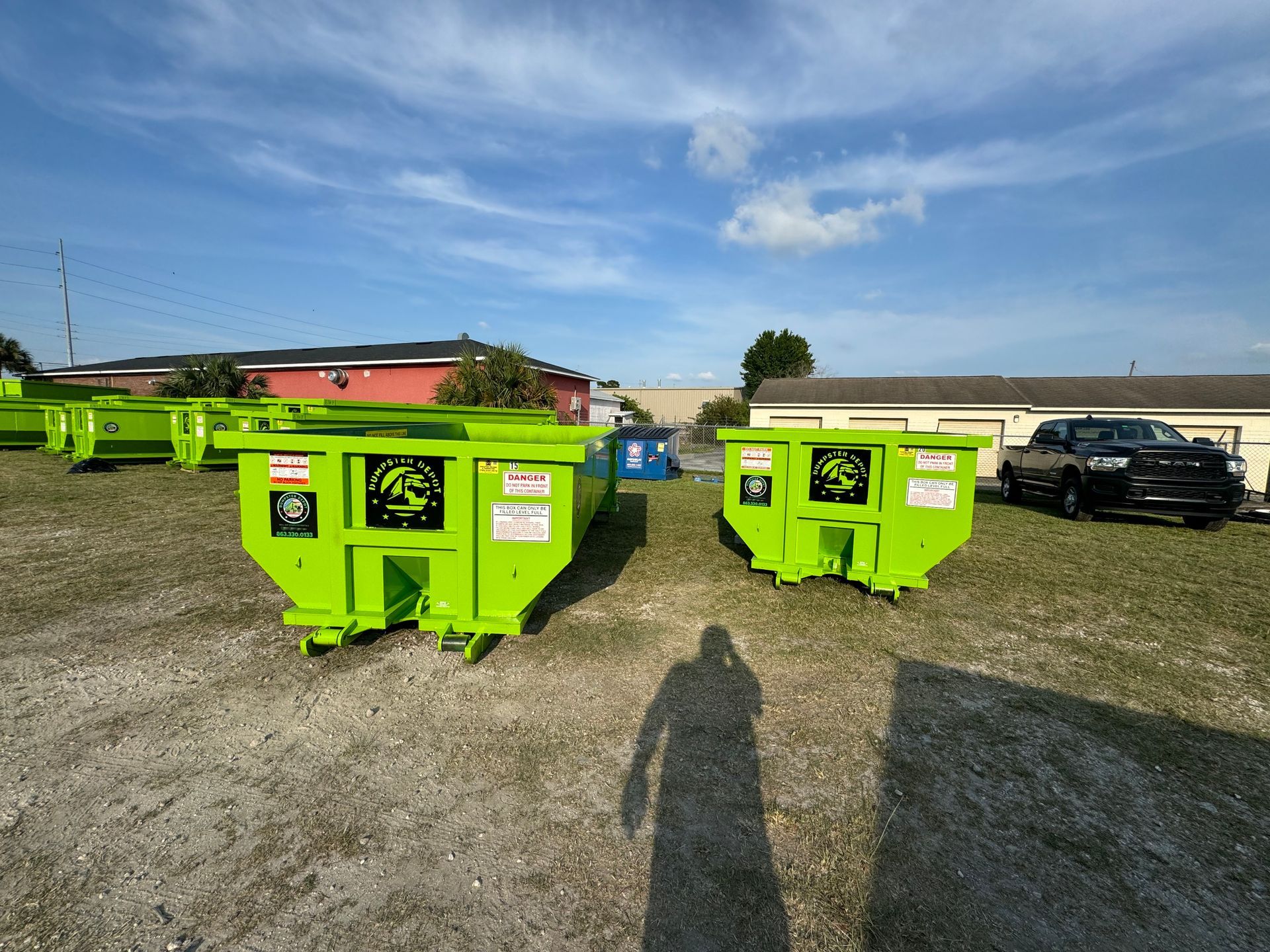 Bright green dumpsters in a gravel lot with parked cars under a blue sky