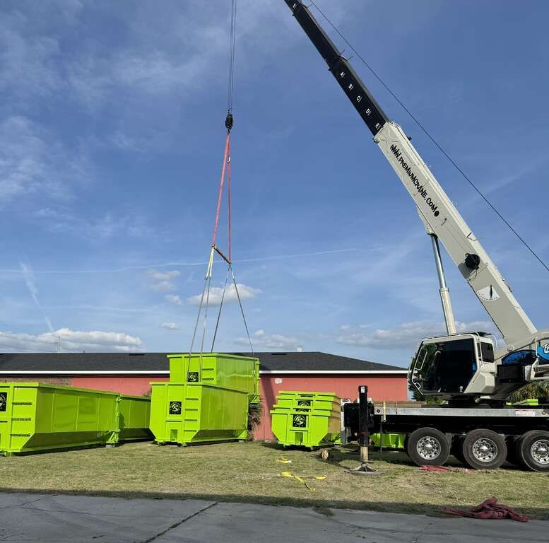 Crane lifting neon green dumpsters in a parking lot under a blue sky