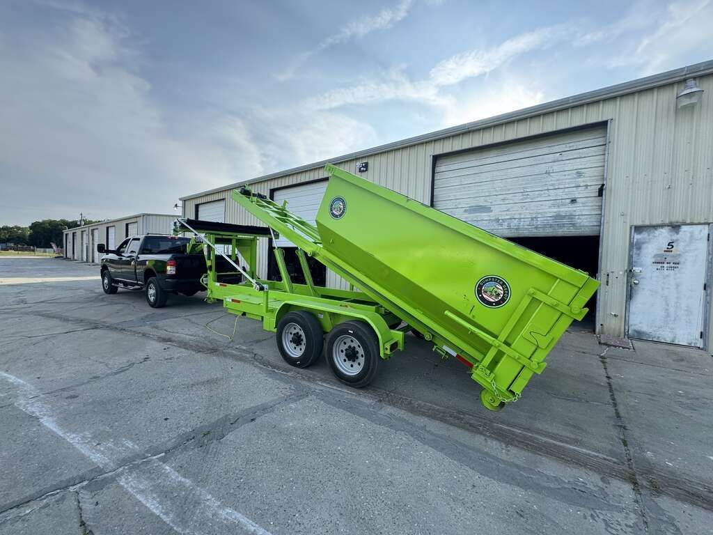 Bright green dump trailer being raised beside a warehouse loading dock, with a pickup truck attached.