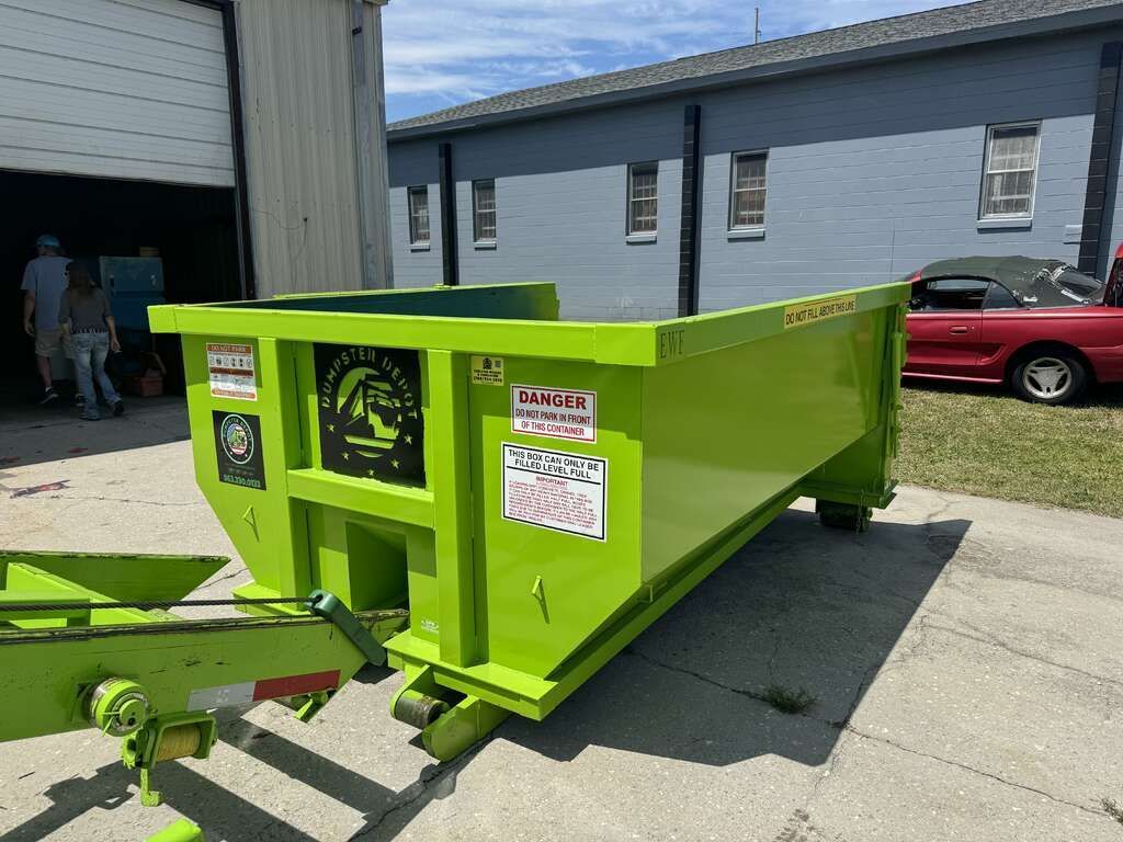 Bright green dumpster trailer parked on a concrete lot beside a building; red car in background.