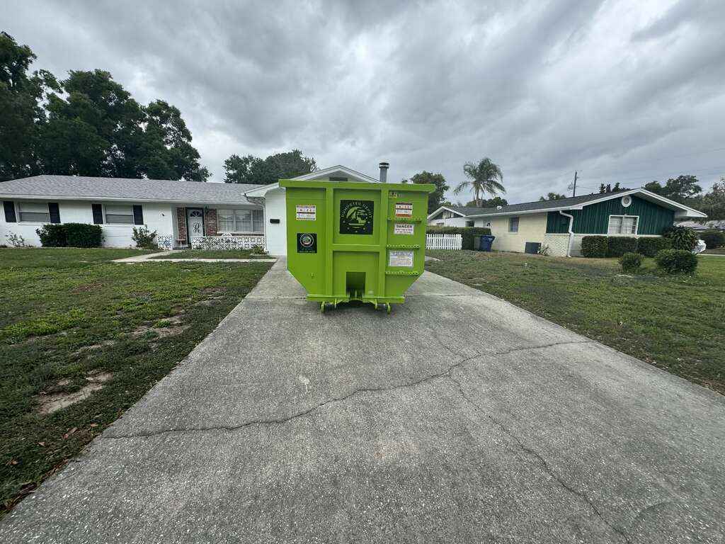 Bright green dumpster on a cracked driveway in a suburban neighborhood under cloudy skies