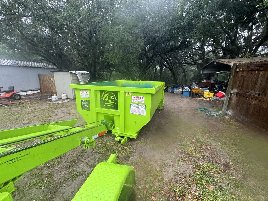 Bright green dumpster trailer in a muddy yard under large trees, with buildings and scattered equipment nearby