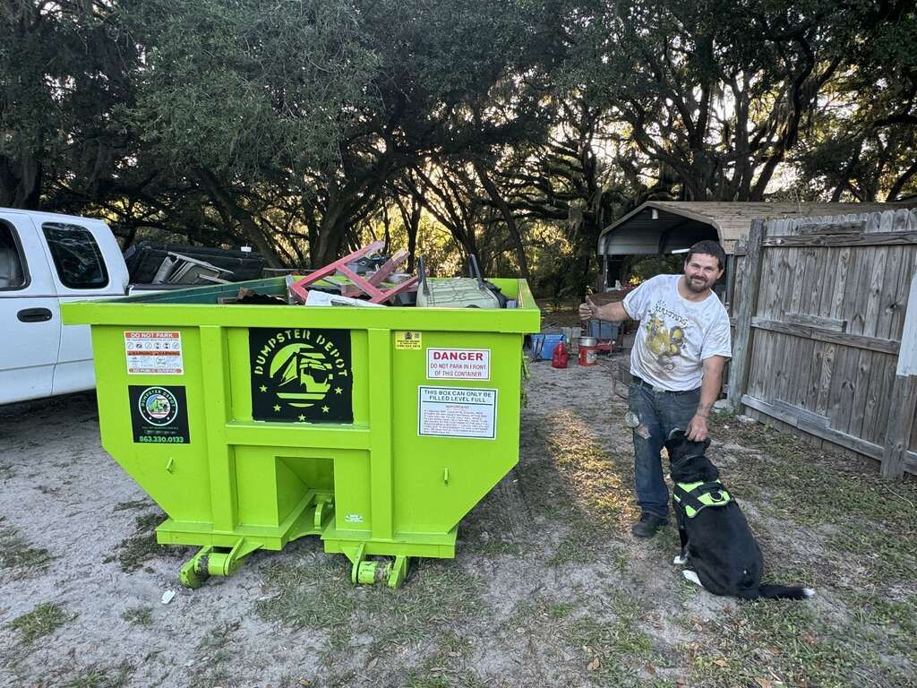 Bright green dumpster in a yard with a man and a black dog beside it, near a white truck and wooden fence.