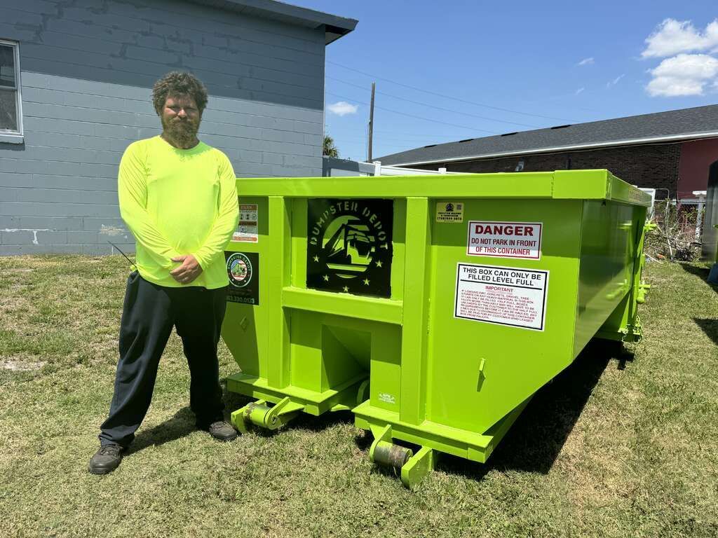 Person standing beside a bright green industrial dumpster outdoors beside a gray building.
