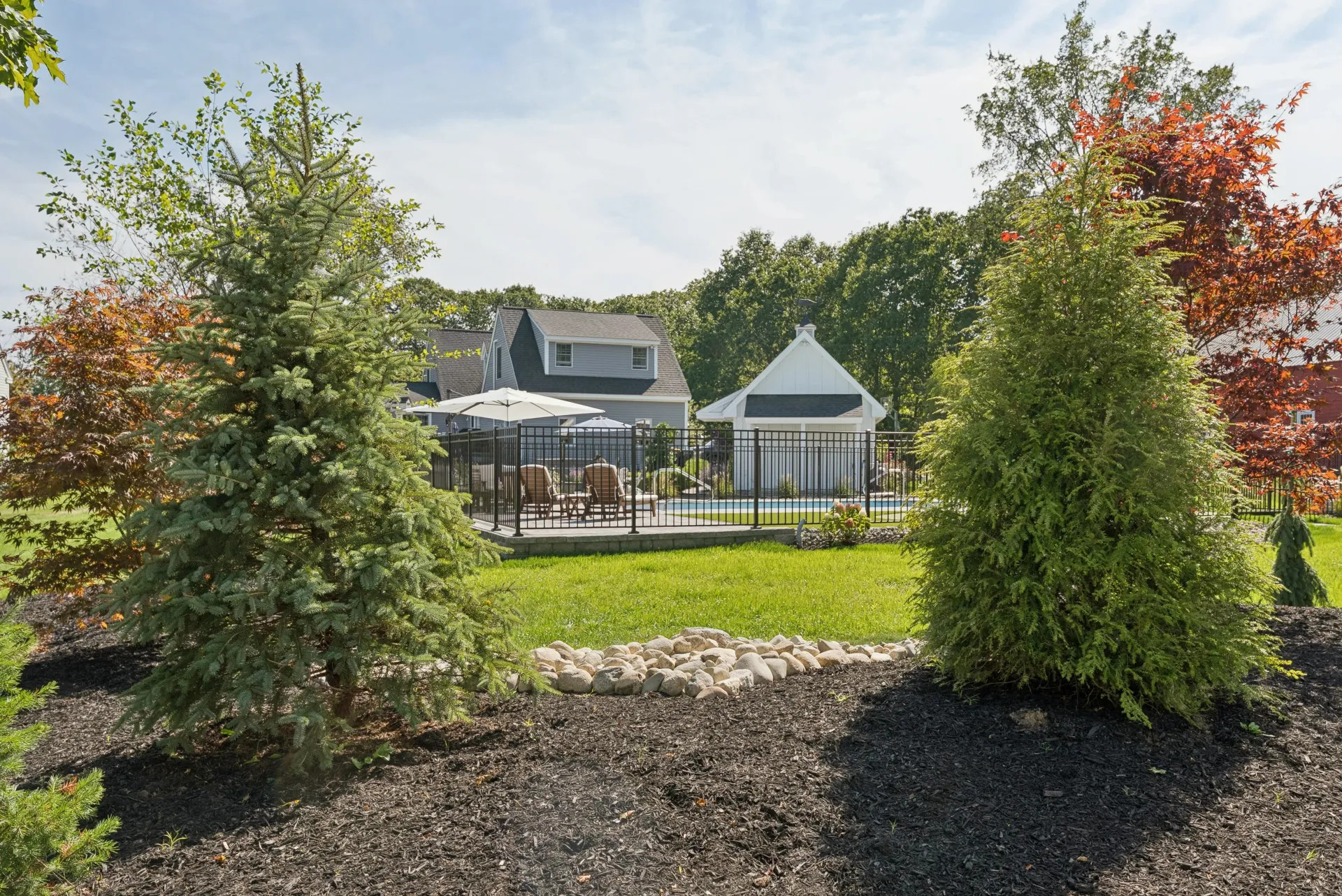 Backyard with pool, patio, and house, surrounded by trees and greenery under a sunny sky.