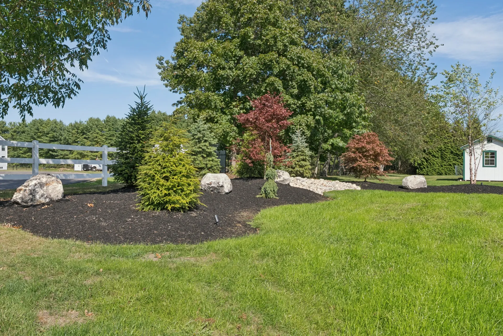 Landscaped yard with black mulch, green grass, trees, rocks, and a white fence against a blue sky.