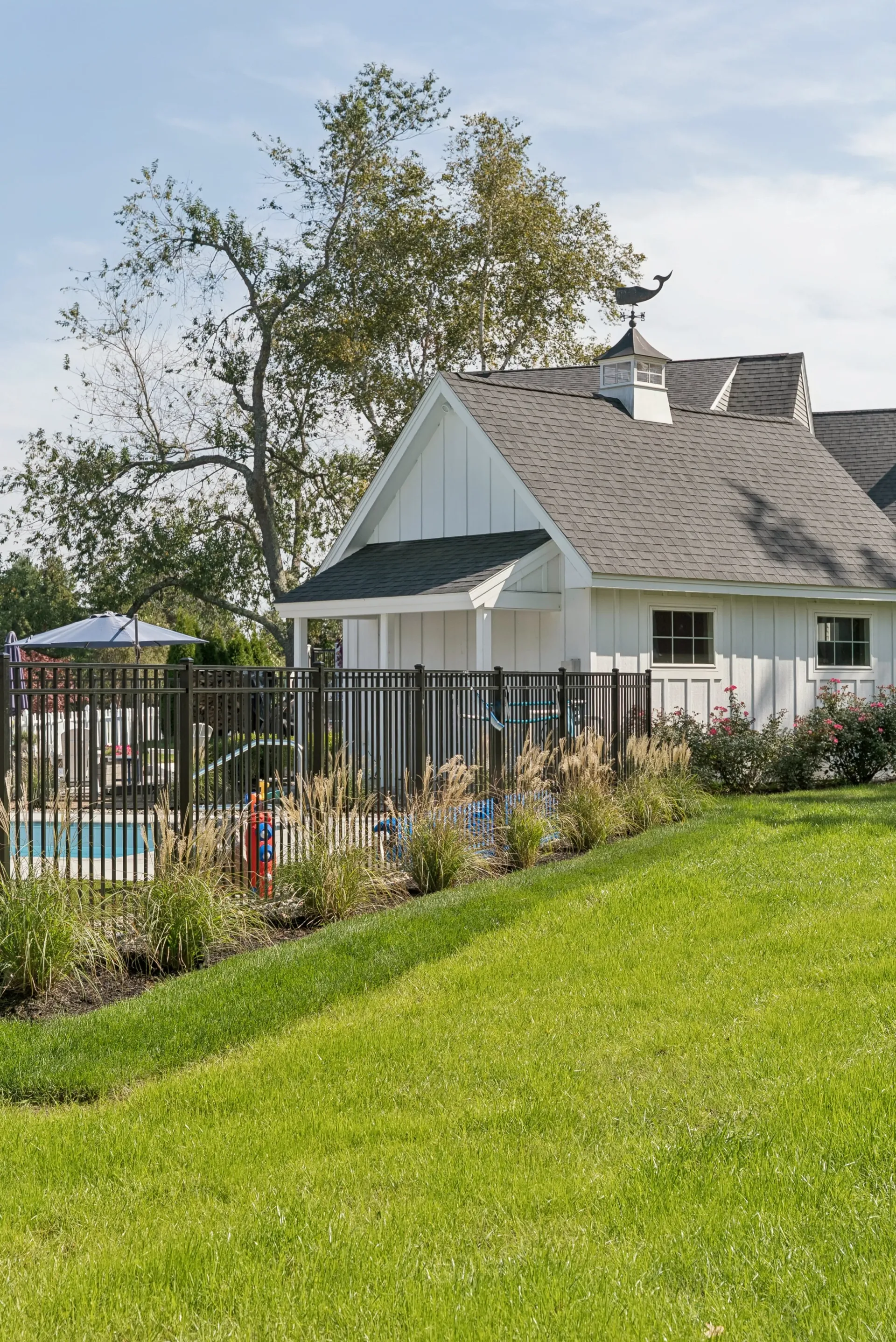 White building with dark roof near a pool, behind a black fence, on a grassy hill.
