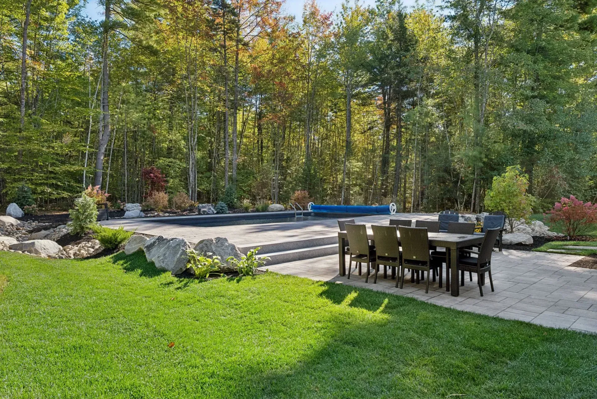 Backyard with pool, patio, and dining table, surrounded by trees.