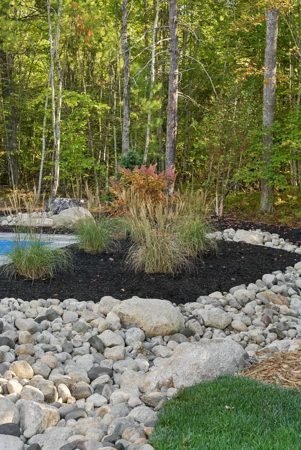 A landscaped garden with a rock bed, mulch, and colorful bushes in front of a forest.
