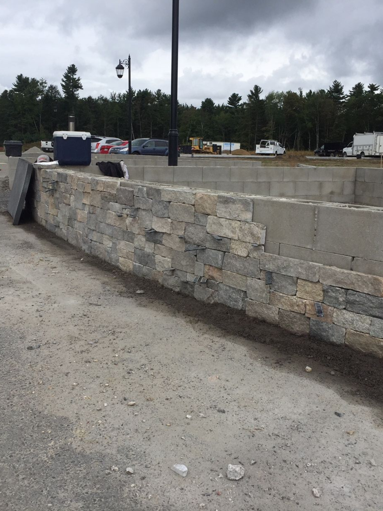 Stone retaining wall beside a gravel lot with parked cars and trees under a cloudy sky