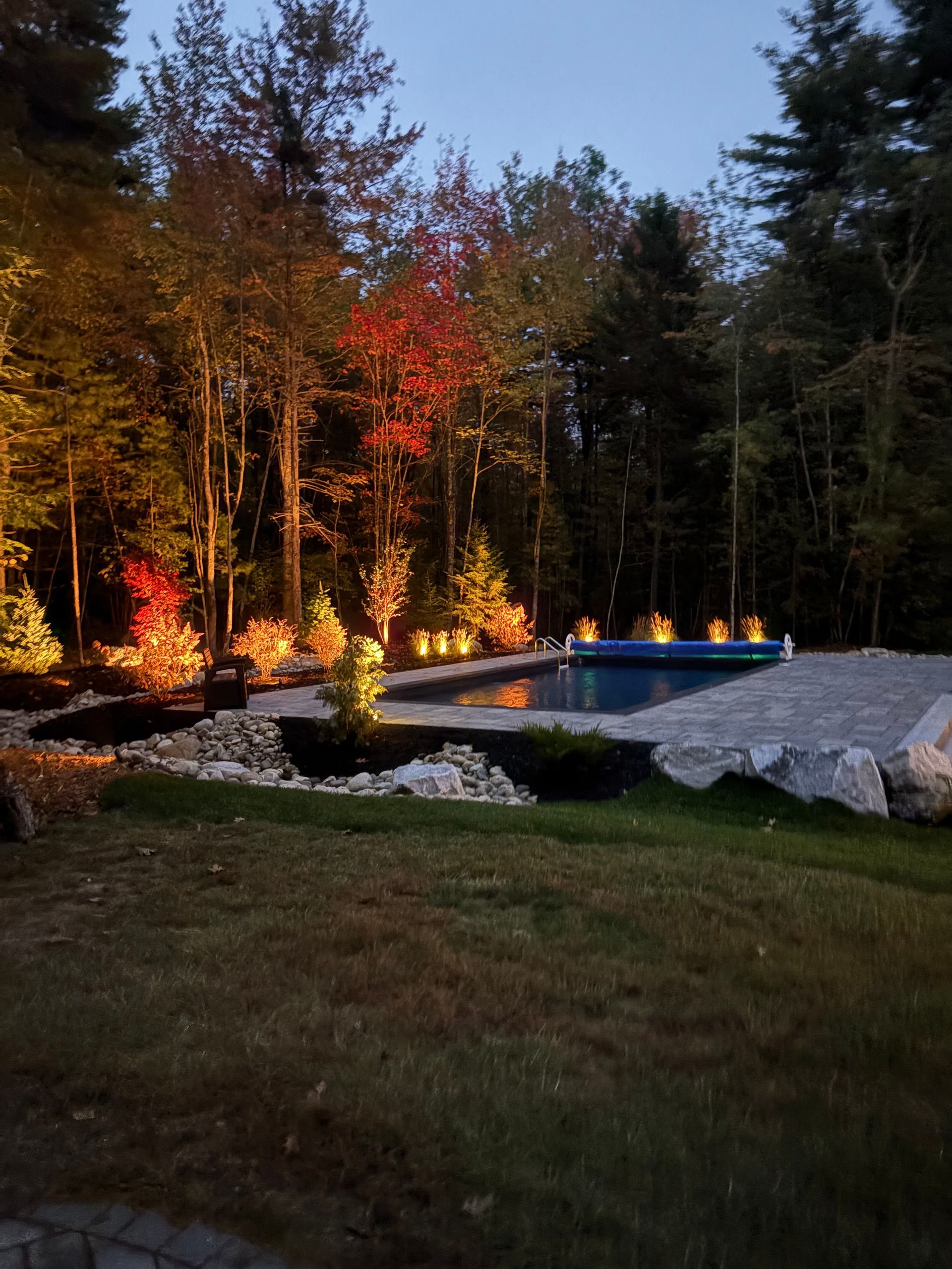 Night view of a pool surrounded by trees, illuminated with spotlights. Fall foliage adds warm colors.
