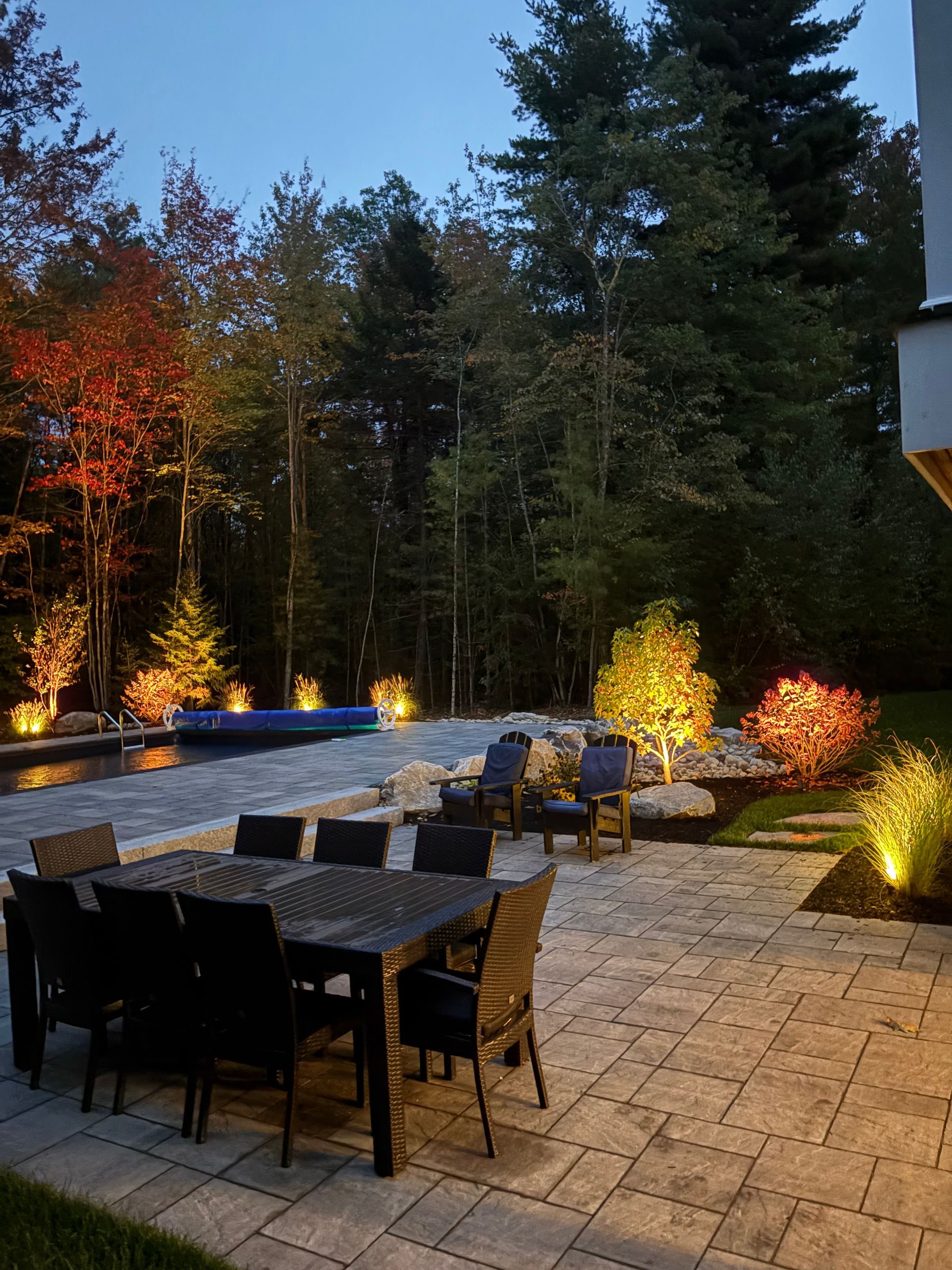 Patio with lit trees, dining set, chairs, and pool at dusk.