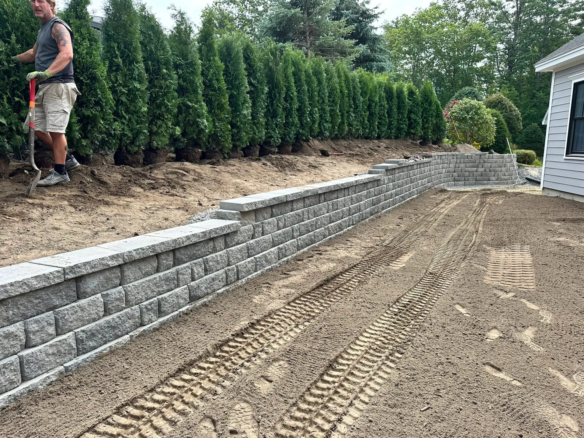 A landscaper trims hedges next to a retaining wall and dirt, near a house.