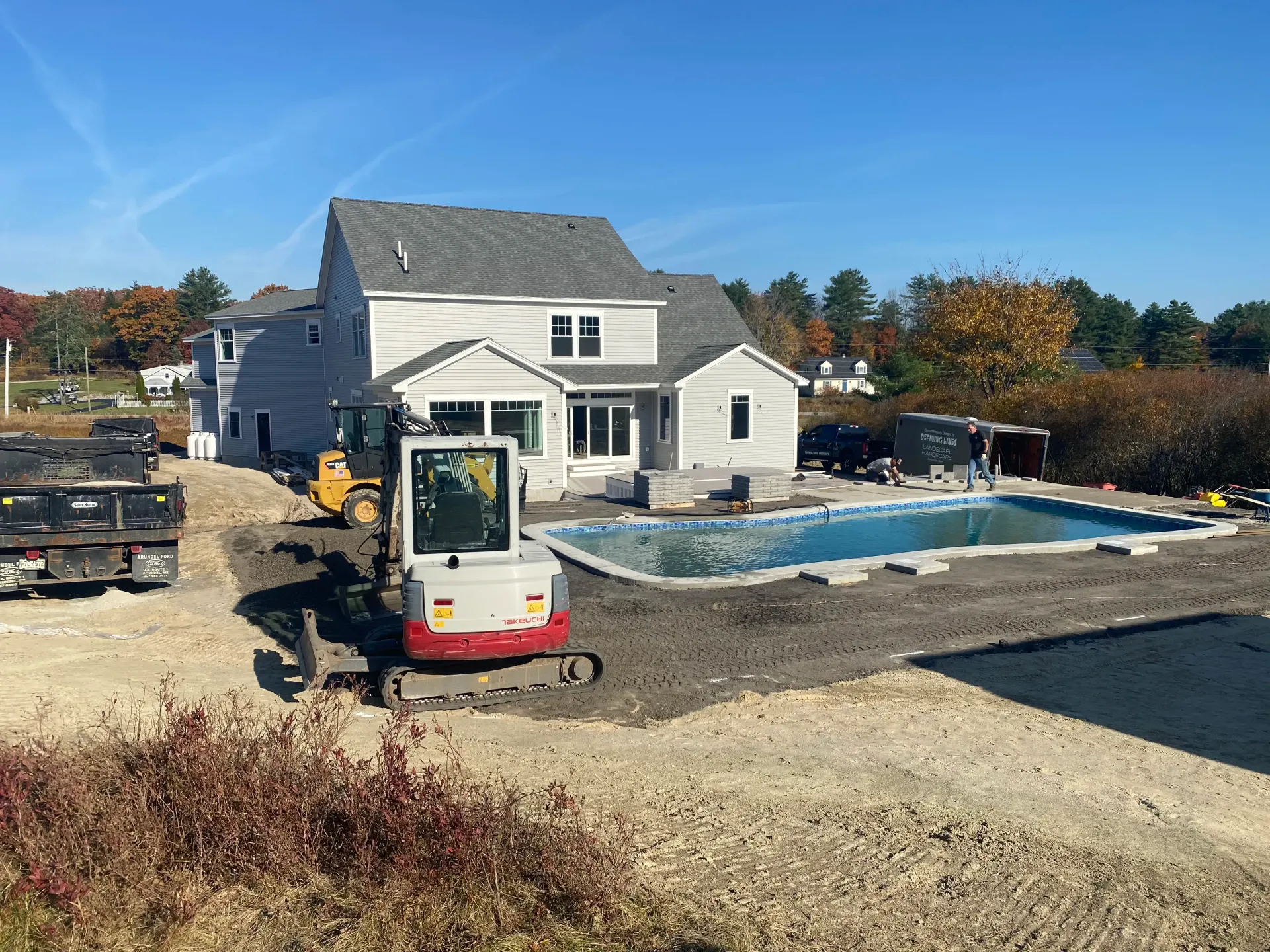 Construction site with house, pool, and machinery. Excavator and dump trucks. Blue sky.