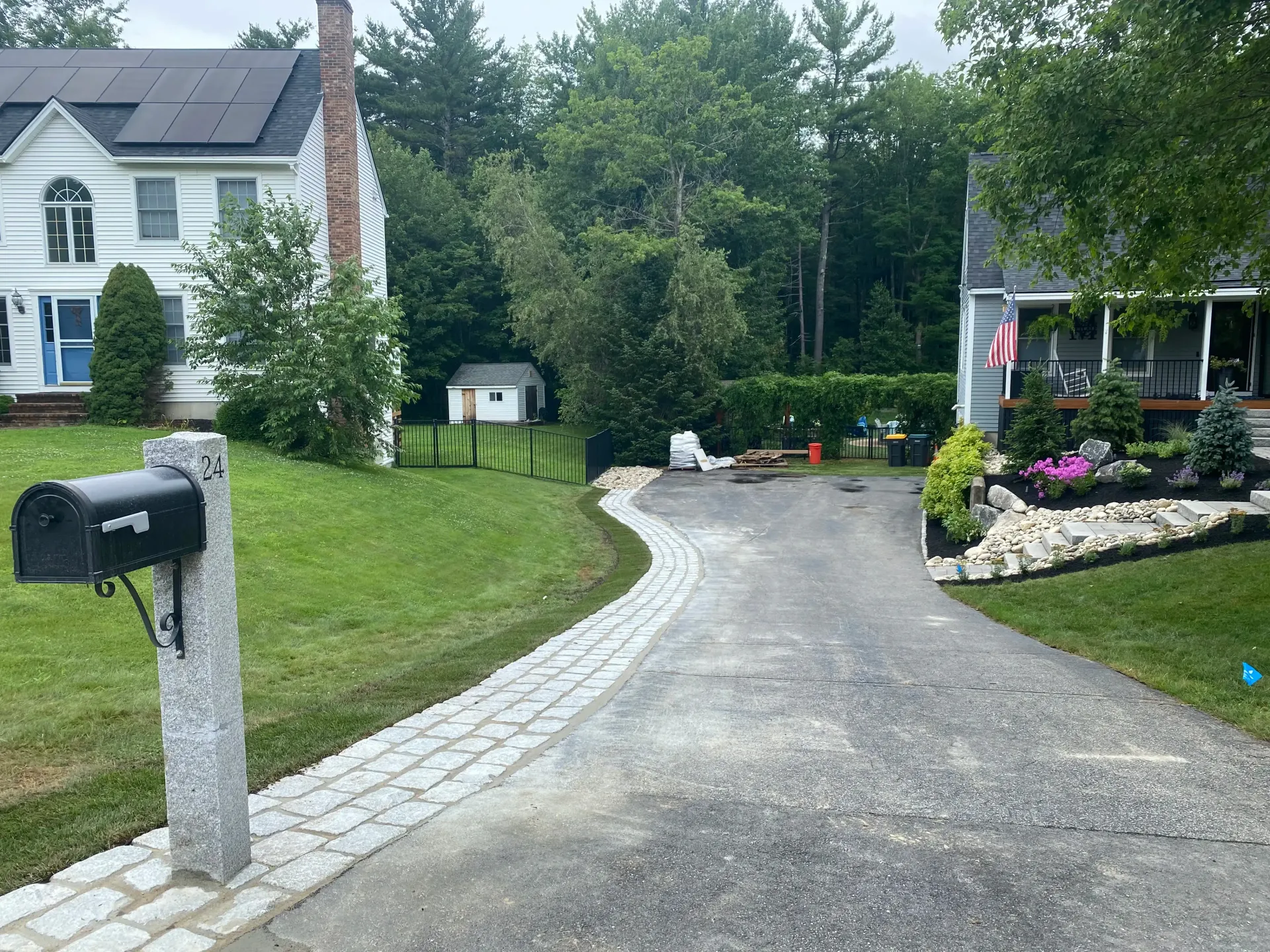 Asphalt driveway curves between two houses. Granite mailbox in foreground, garden bed, and trees behind.