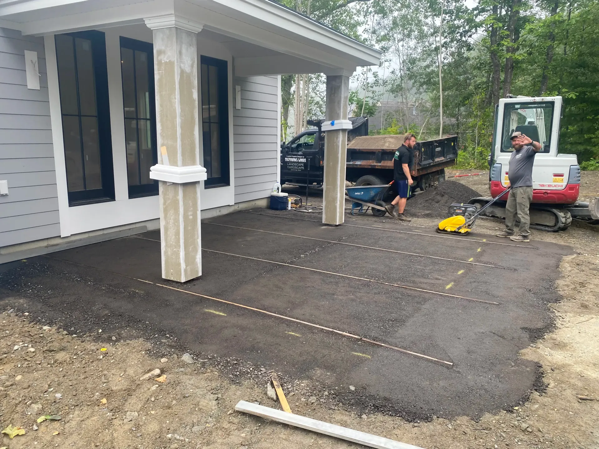 Workers paving a patio next to a house with columns. Heavy machinery and a truck are present.