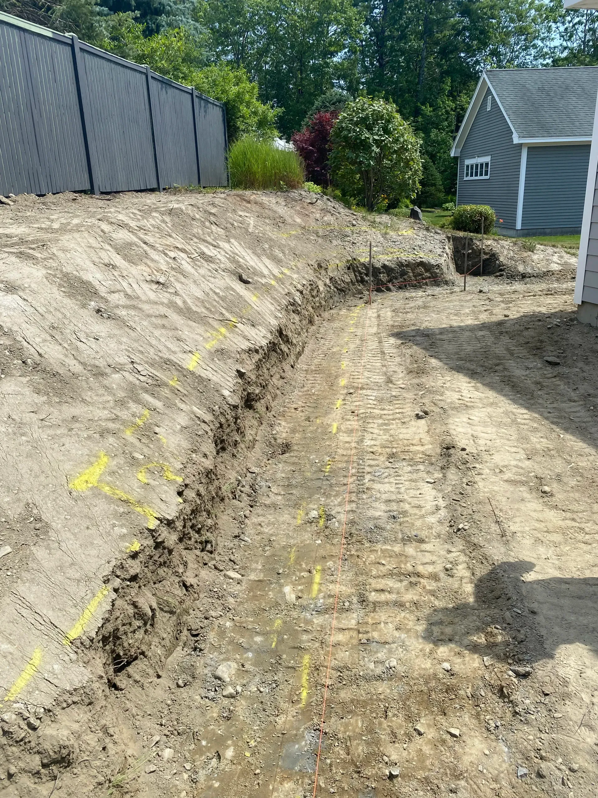 A trench dug in dirt next to a gray fence and a house, with yellow markings.