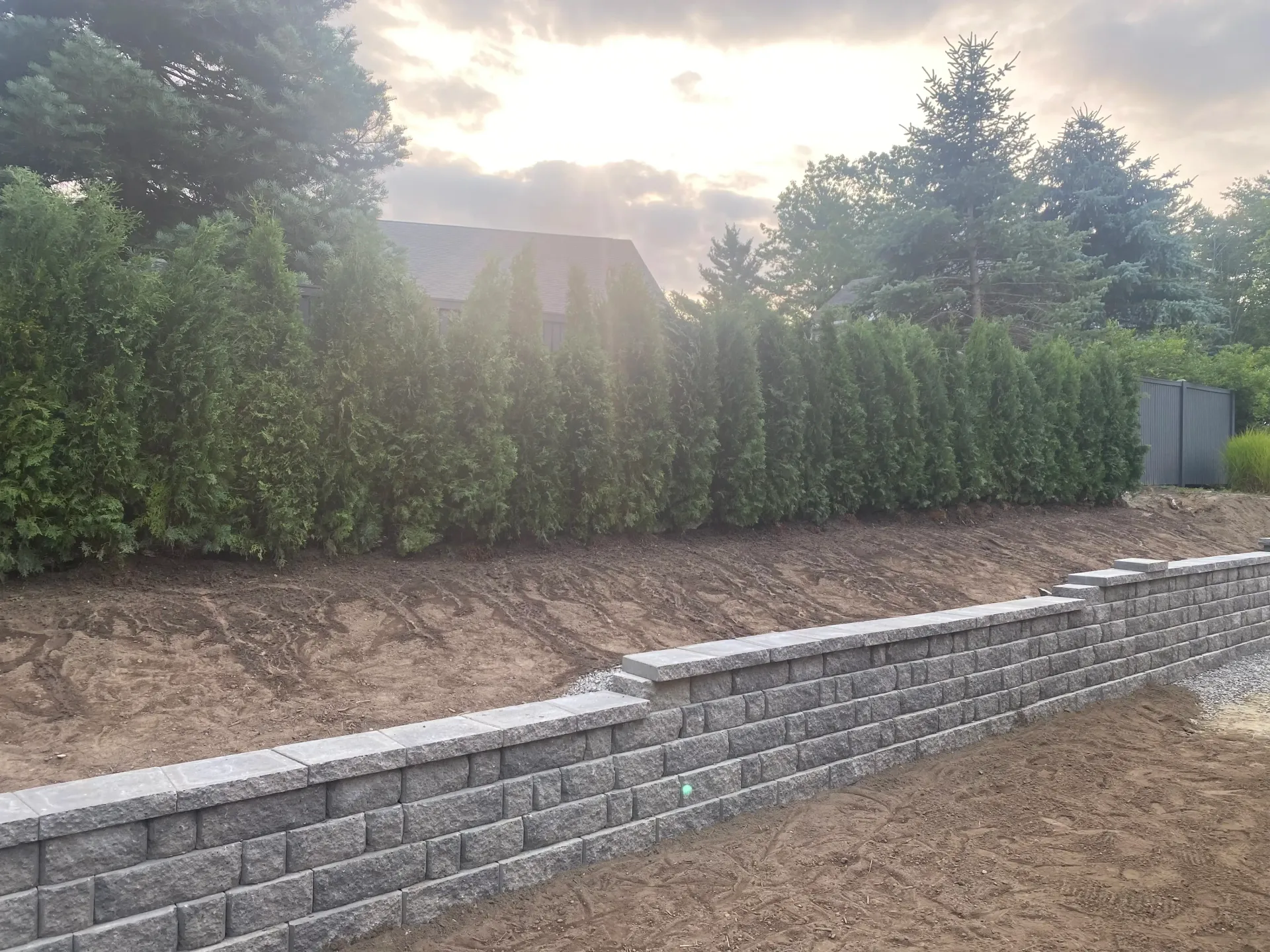 A stone retaining wall in front of a row of evergreen trees and a cloudy sky.