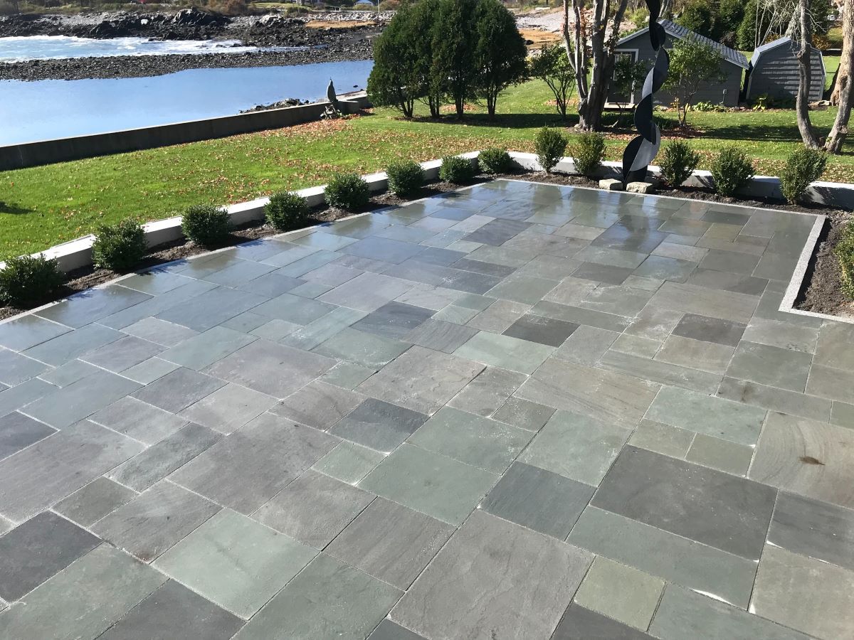 Stone patio beside a lawn and water, lined with shrubs and trees in a landscaped yard