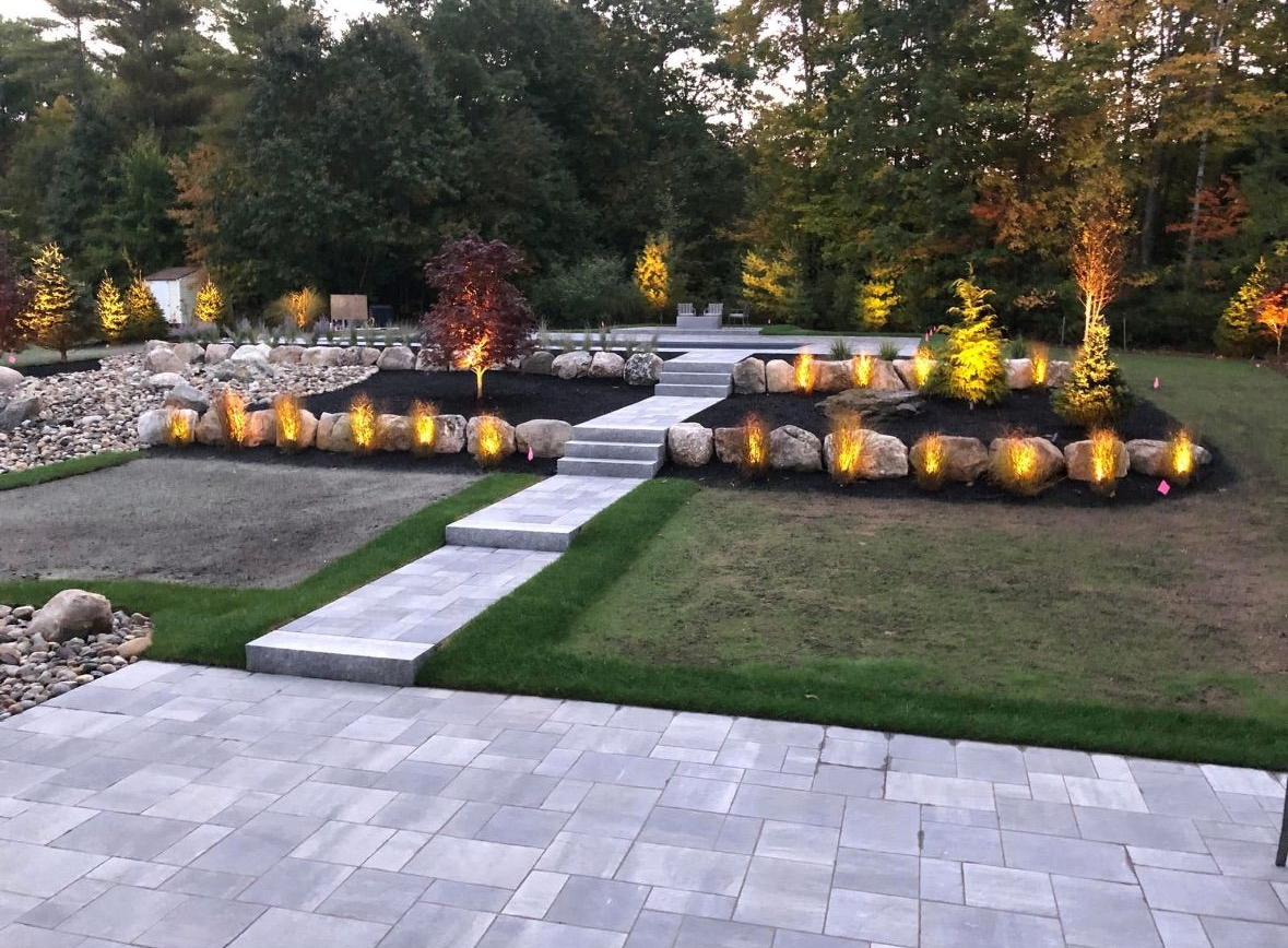 Backyard landscape with stone patio, steps, illuminated rock beds, and trees.