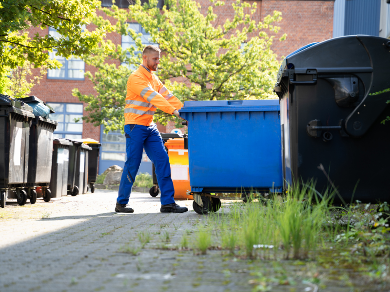 Worker in orange vest pushing a blue waste bin towards a dumpster in an alley.