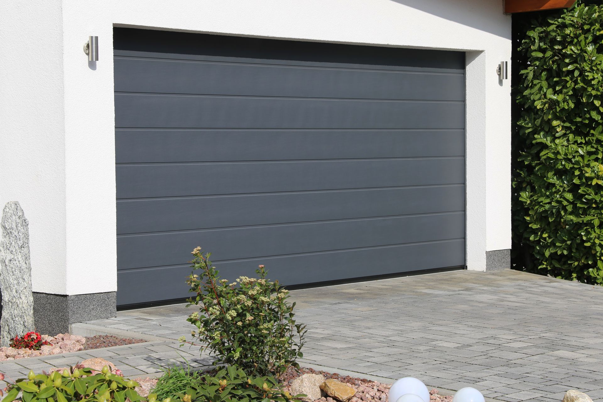 A dark gray, horizontal-slatted sectional garage door installed in a white house, with a paved driveway in front.