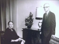 Two people in a living room, one seated by a curtain and one standing beside a lamp and cabinet.