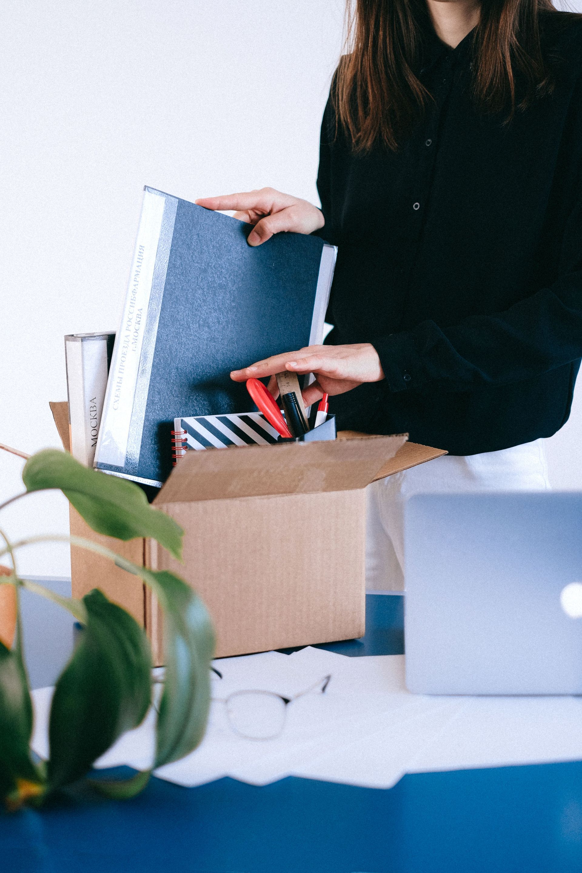 Employee packing blue folders into a cardboard box at a desk with a laptop and plant