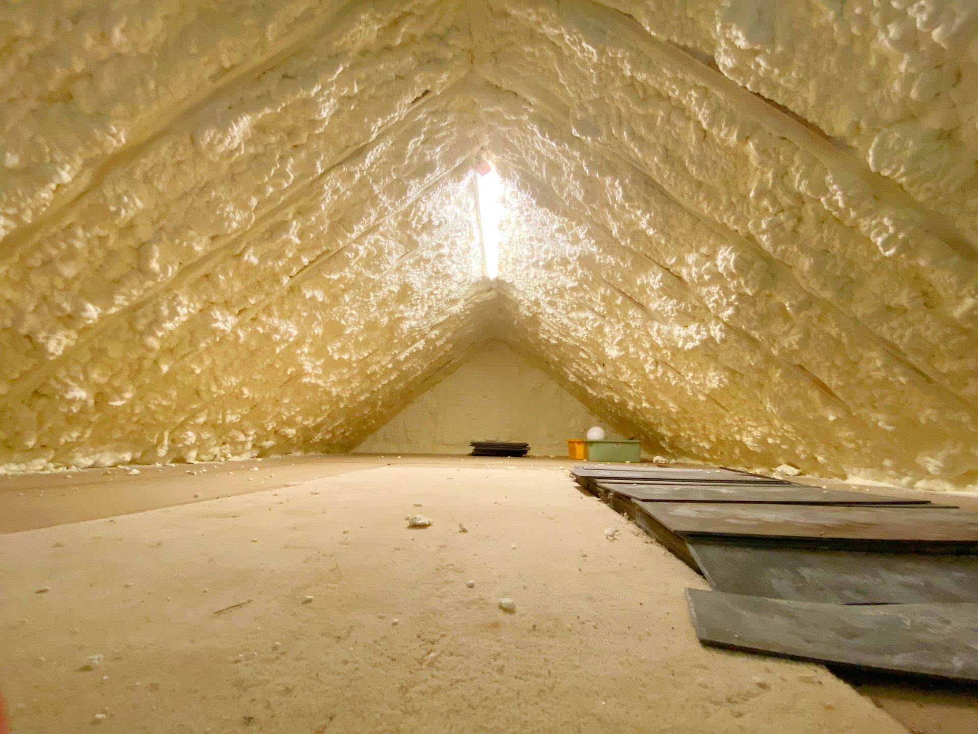 Attic with spray-foam insulation and a wooden plank walkway leading toward a bright opening