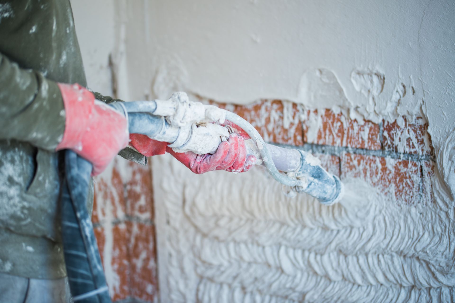 Worker scraping plaster from a wall with a putty knife, revealing brick beneath