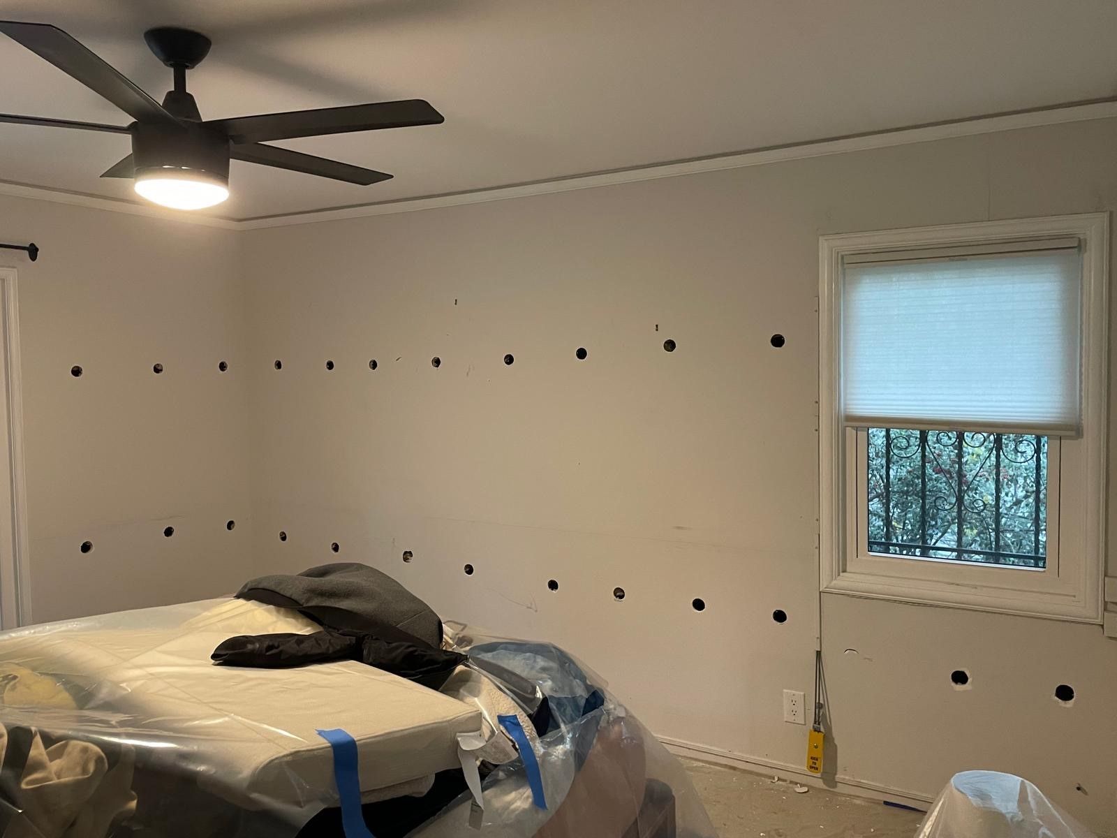 Partially renovated bedroom with bed, ceiling fan, wall holes, and a window with blinds
