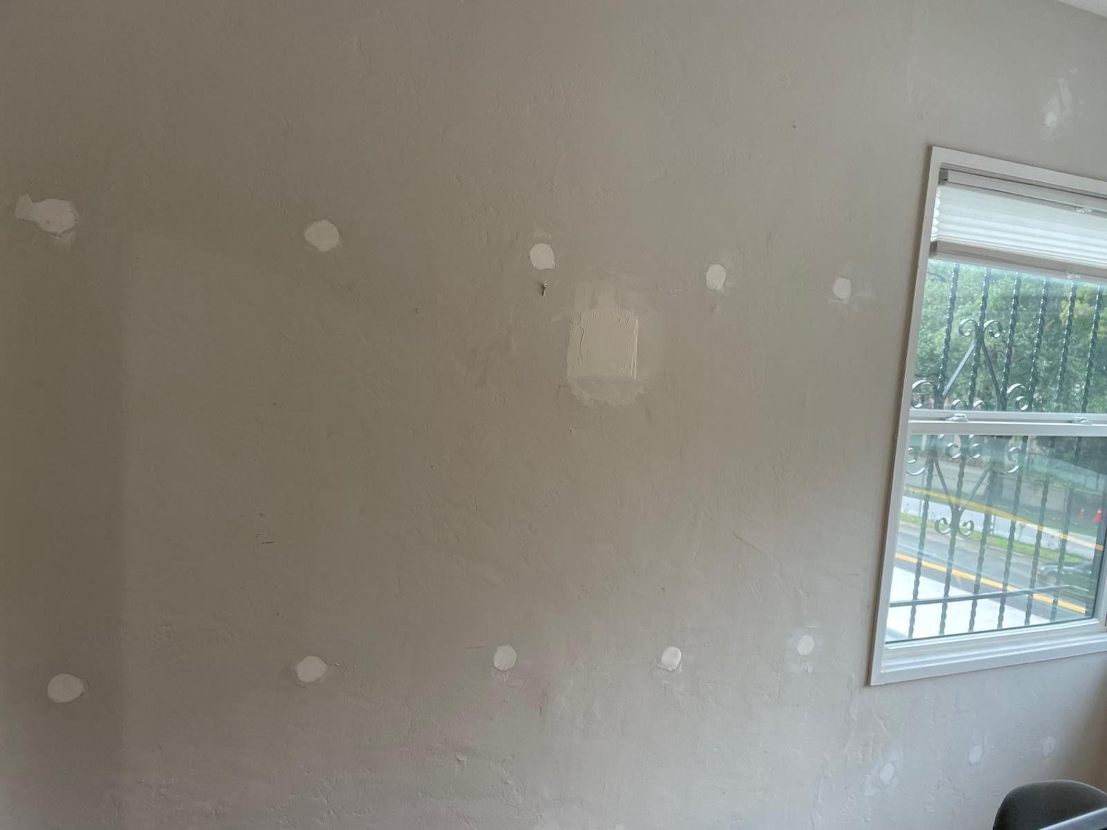 Empty beige wall with patchy repairs and a small window showing a barred view outside.