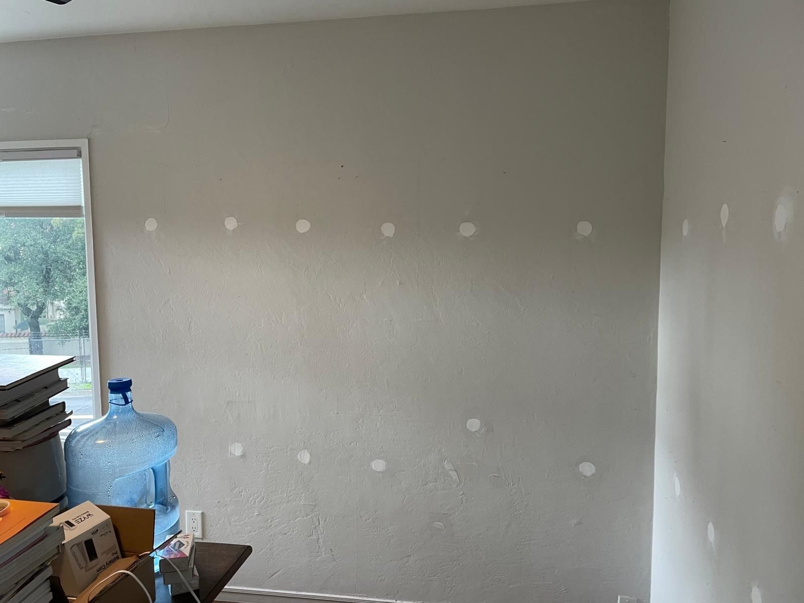 Empty room with light gray walls and white patch marks, window at left, and stacked items in the corner