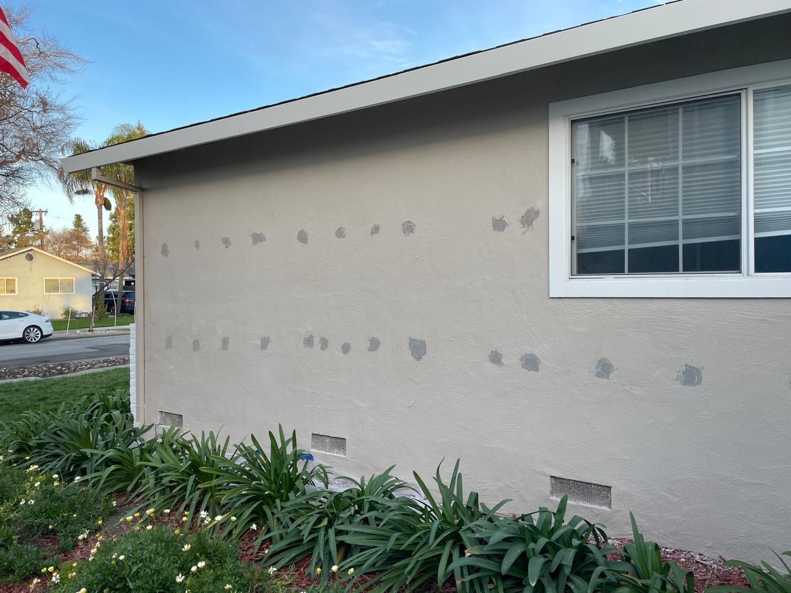 Beige house exterior wall with multiple patch marks, window, and shrubs along the base.