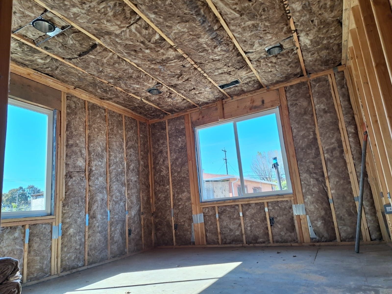 Unfinished room with exposed insulation, wooden framing, and two windows letting in sunlight.