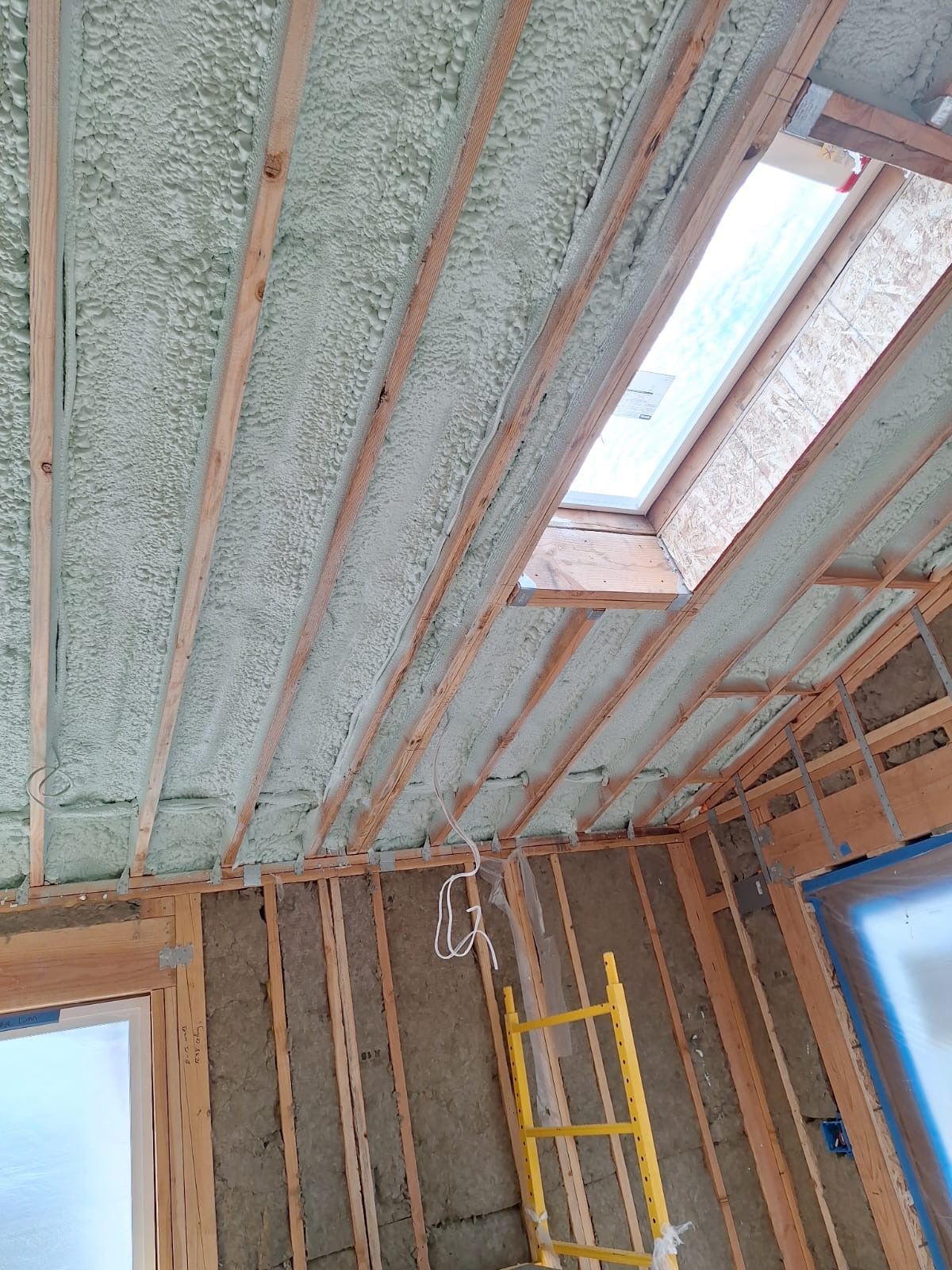 Insulated attic ceiling under construction with exposed wooden rafters, skylight, and a yellow ladder.