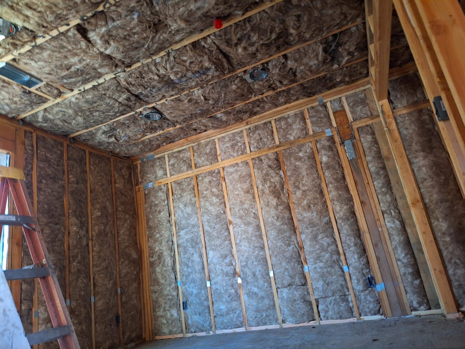 Unfinished room with exposed insulation, wooden studs, and a ladder leaning against the left wall.