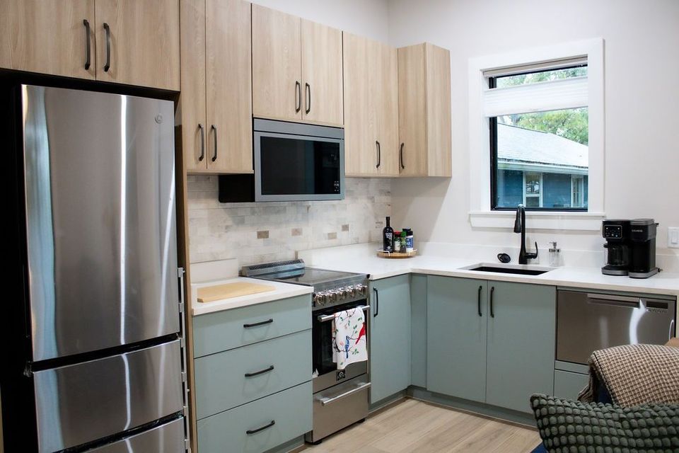 Modern kitchen with stainless steel appliances, light wood and teal cabinets, white countertops, and small window.