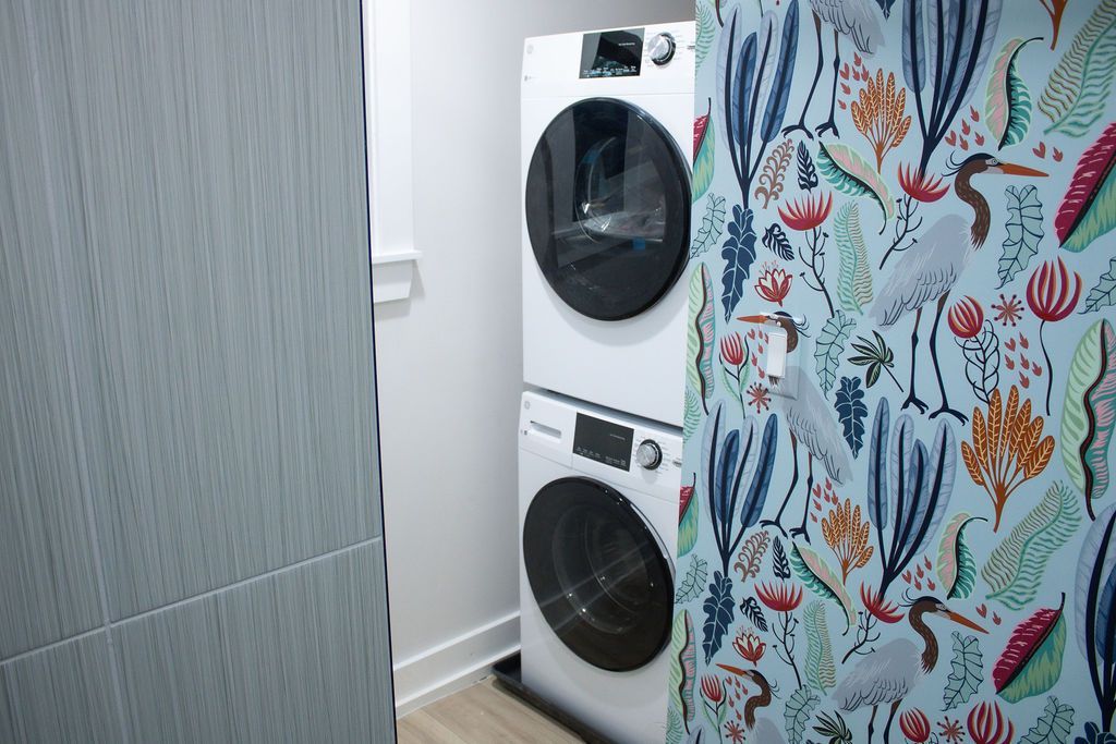 Stacked white washer and dryer in a small laundry area, next to a patterned wall.