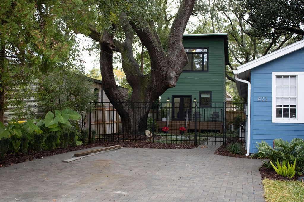 Green house behind a large tree, blue house to the right, and brick driveway.