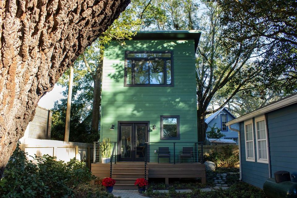 Green two-story house with a deck and sliding glass door, framed by trees and plants.