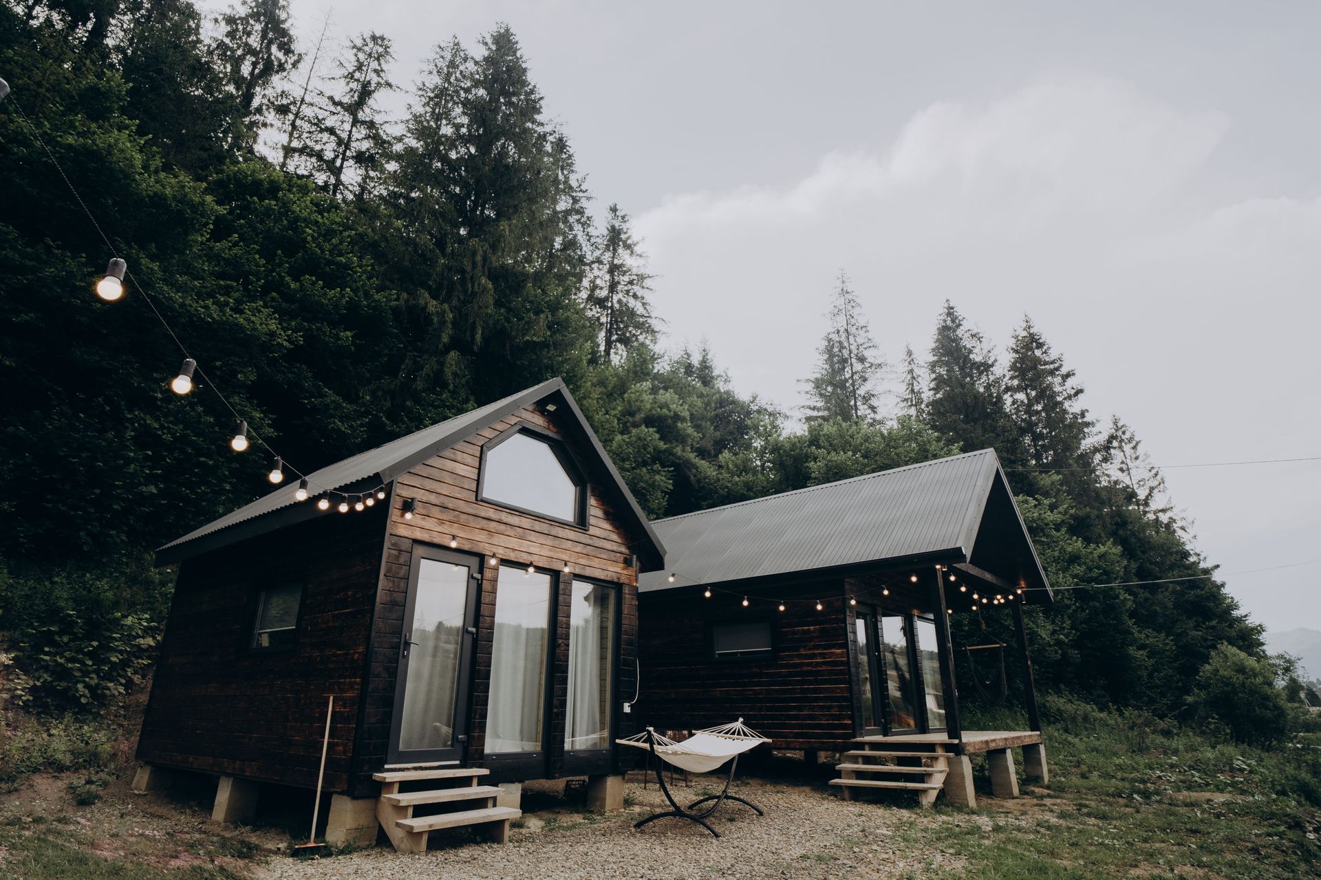 Two rustic cabins in a wooded clearing with string lights and picnic tables under an overcast sky