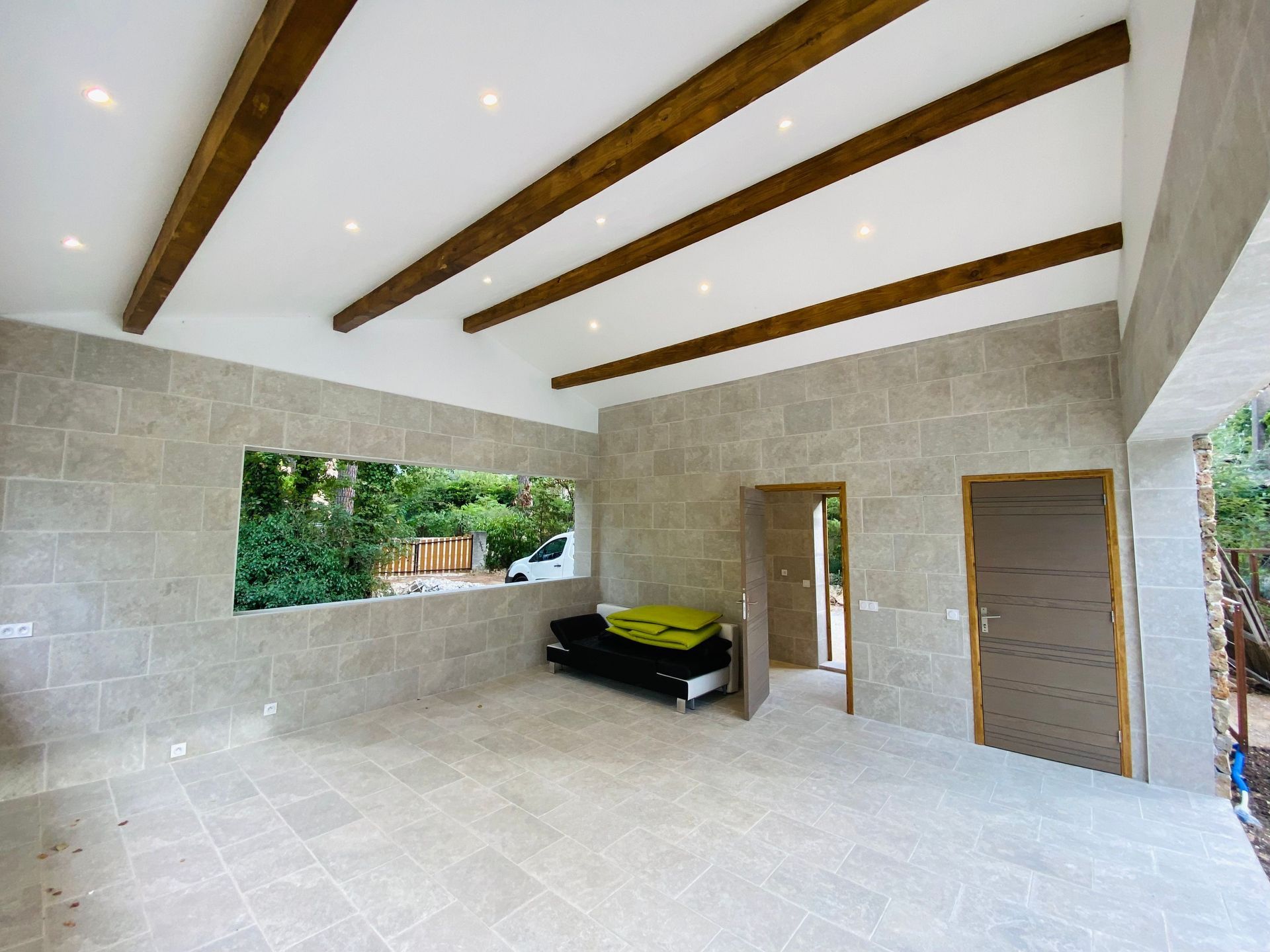 Modern open-air room with stone walls, white ceiling, exposed beams, and large windows overlooking greenery