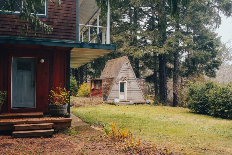 Rustic cabin and tiny A-frame cottage in a wooded yard with tall trees and grass