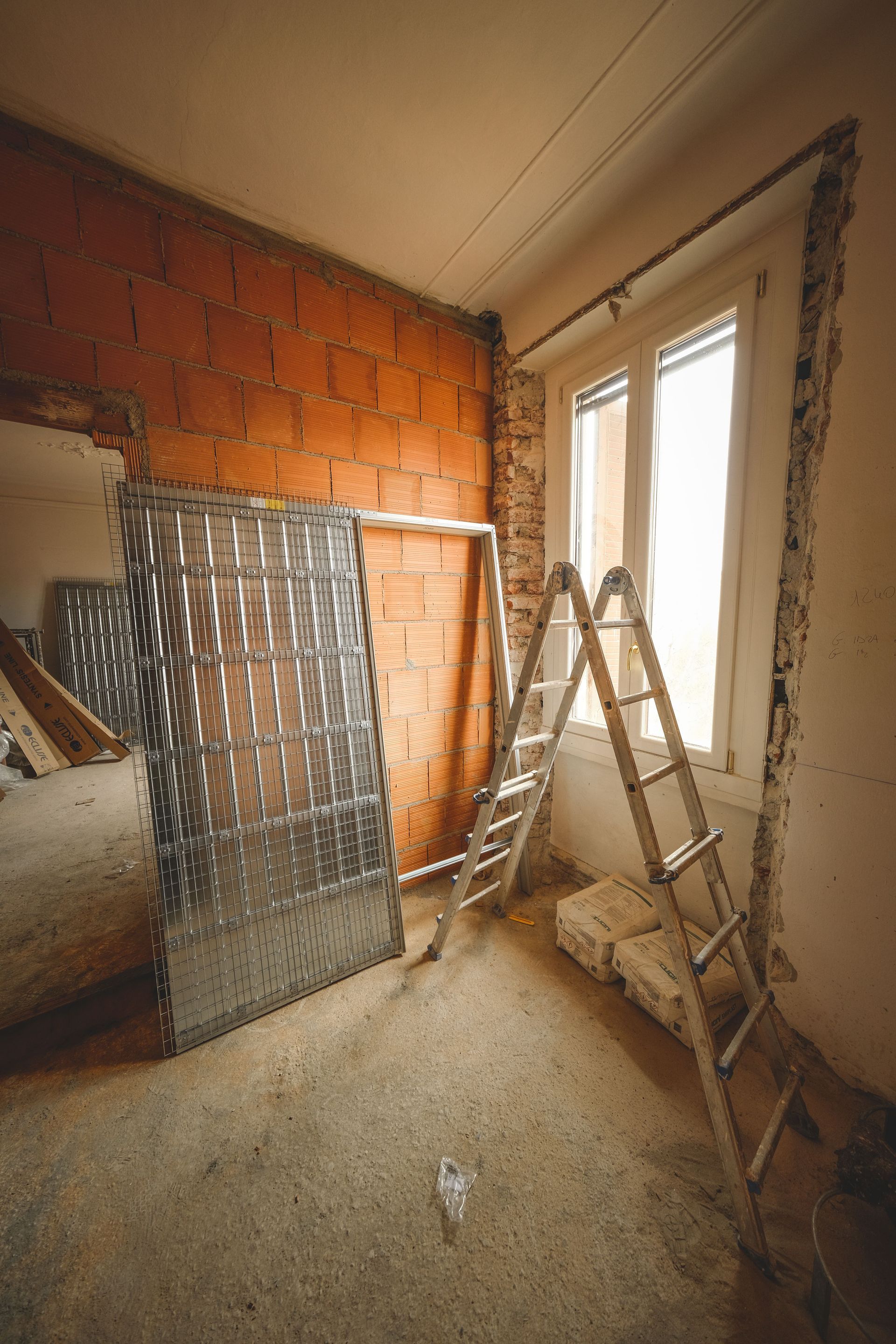 Renovation site with red brick wall, ladder, and reflective insulation leaning in a dusty room near a window.