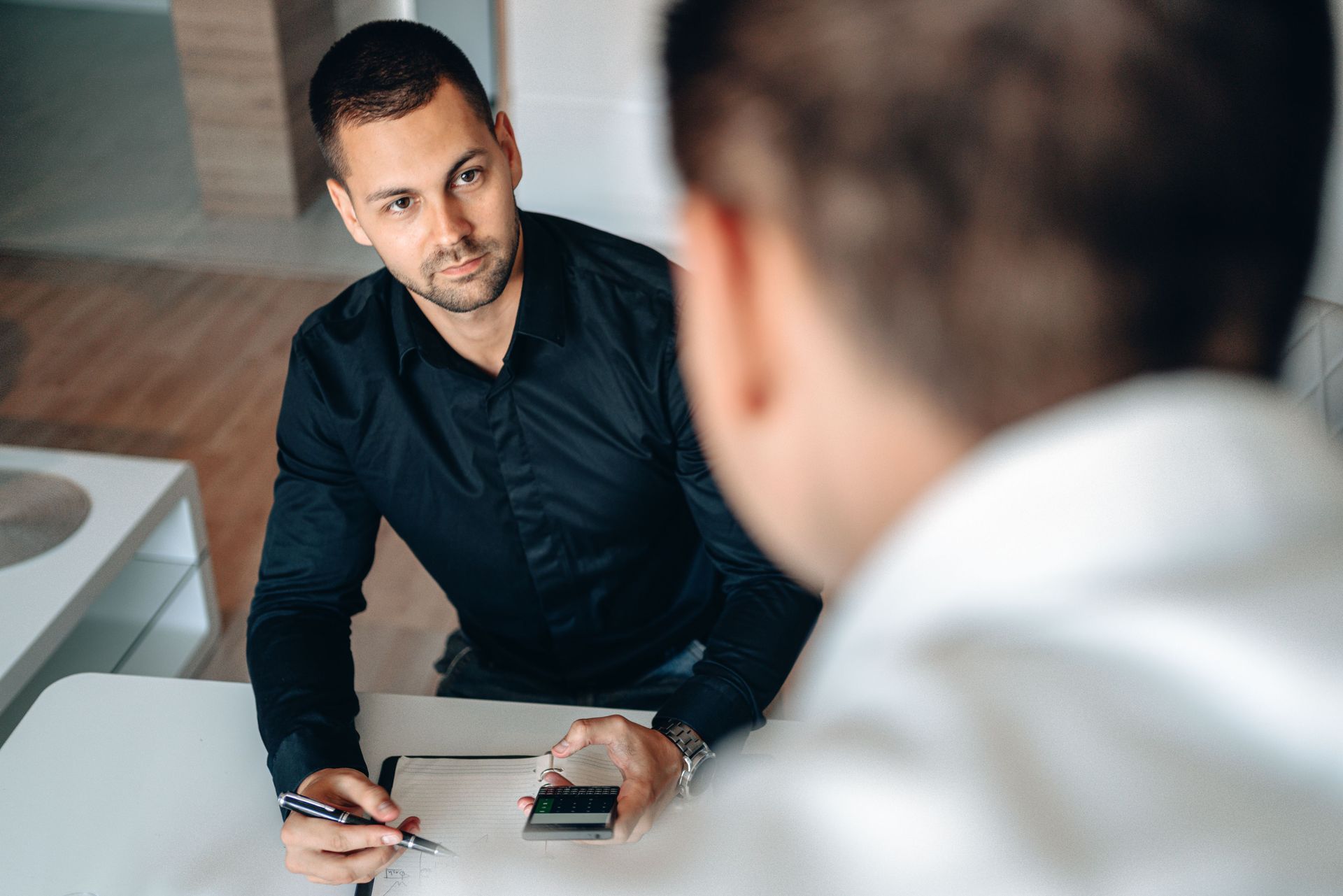 Two people in a meeting, one taking notes while speaking across a desk