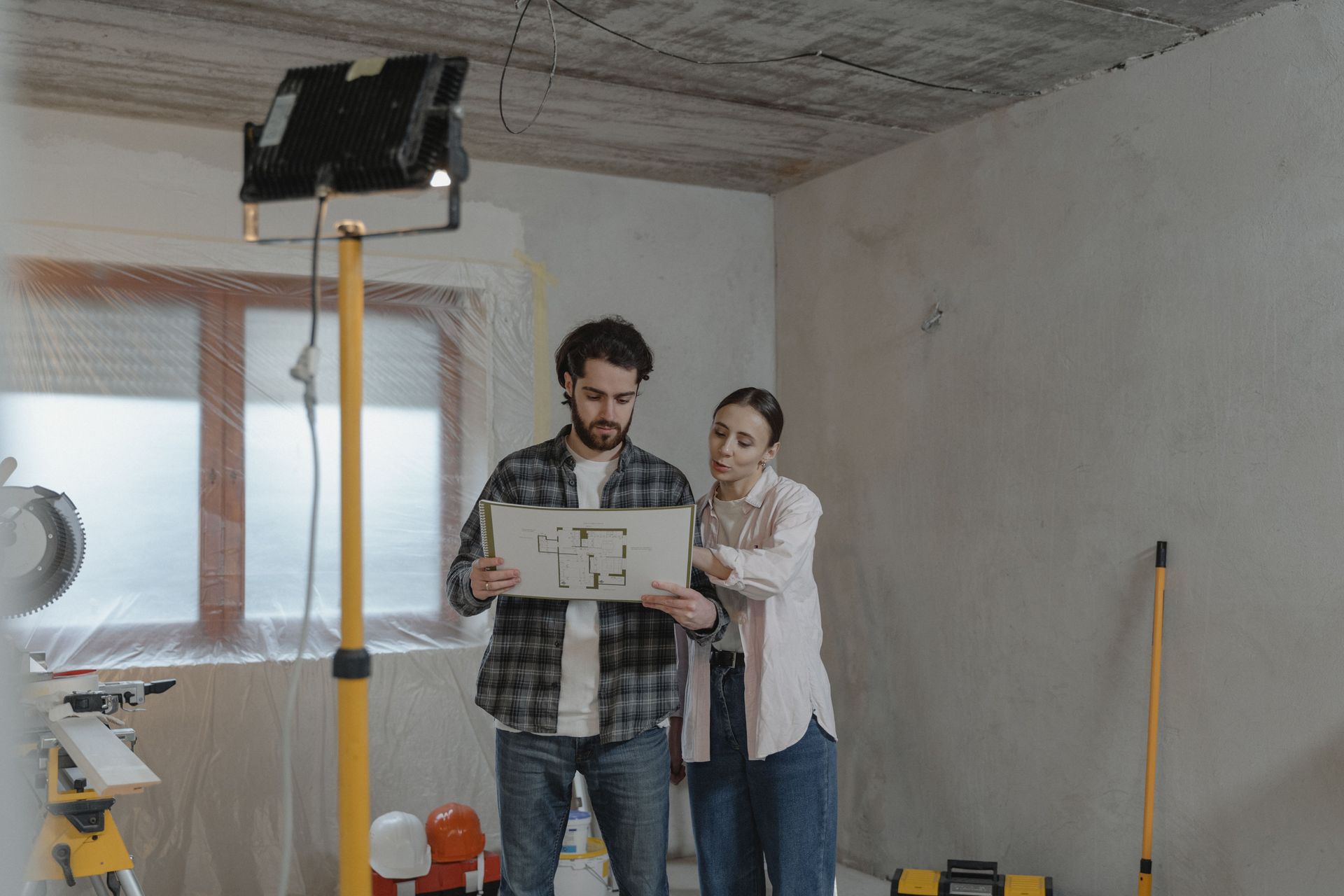 Two people reviewing a laptop in an unfinished room with a work light and tools nearby